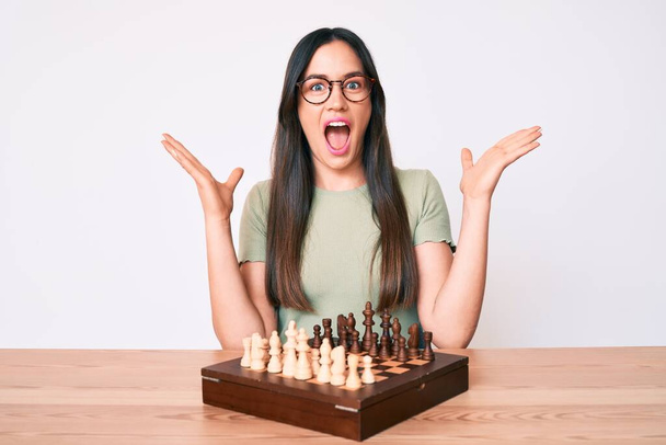 Genç beyaz kadın masada oturmuş satranç oynuyor zaferi mutlu bir gülümsemeyle kutluyor ve kazanan el kaldırıyor.  - Fotoğraf, Görsel