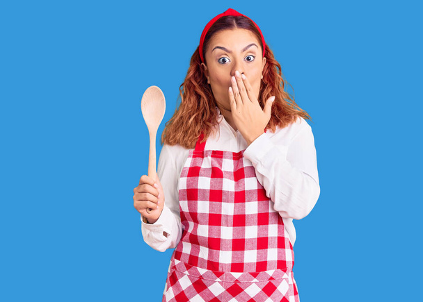 Young latin woman wearing apron holding wooden spoon covering mouth with hand, shocked and afraid for mistake. surprised expression  - Photo, Image