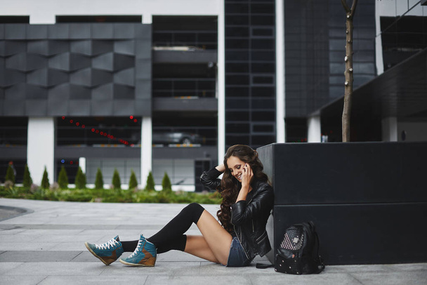 A young woman in denim shorts and leather jacket talking by phone and sitting on the street of a European city on a summer day - Foto, imagen