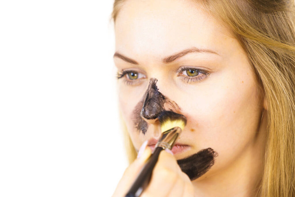 Jonge vrouw wordt toegepast met borstel zuiverende houtskool masker, zwarte cosmetica op haar gezicht. Vrouwelijke verzorging van de vette huid. Schoonheidsbehandeling. Huidverzorging. - Foto, afbeelding