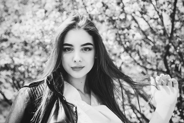 Young beautiful girl with long hair enjoys the beauty of spring nature near the blossoming sakura tree. Girl in a leather jacket in sakura. Black and white photo of a girl. Portrait - Fotografie, Obrázek