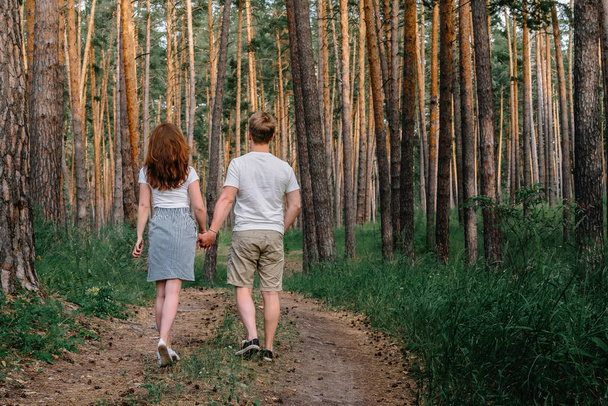  Ein blonder Mann und ein Mädchen mit langen Haaren im Rock gehen Hand in Hand durch einen Kiefernwald - Foto, Bild