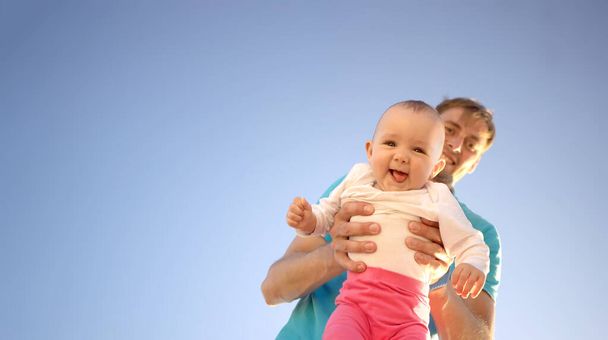 Father holds a small child in his arms on a background of a summer blue sky. Cute baby is smiling. Happy family outdoors. - 写真・画像