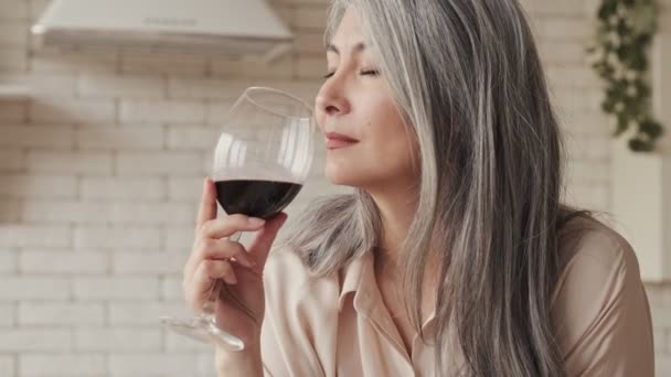 A beautiful mature woman is sitting at the table in the kitchen and holding a glass of red wine - Filmati, video