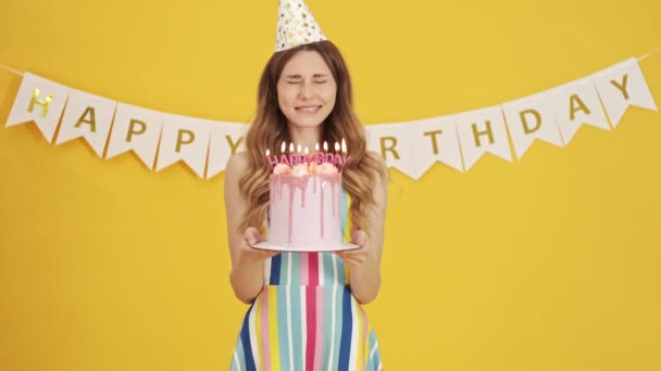 A happy woman in a party cone is making a wish while holding a birthday cake then blowing candles isolated over a yellow background - Video