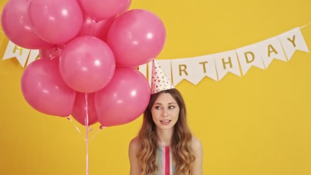 An attractive woman wearing a party cone is being lifted with pink balloons isolated over a yellow background - 映像、動画