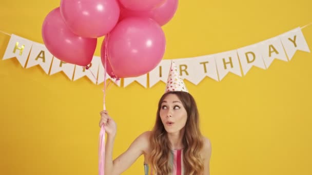 An excited woman wearing a party cone is being lifted with pink balloons isolated over a yellow background - Imágenes, Vídeo