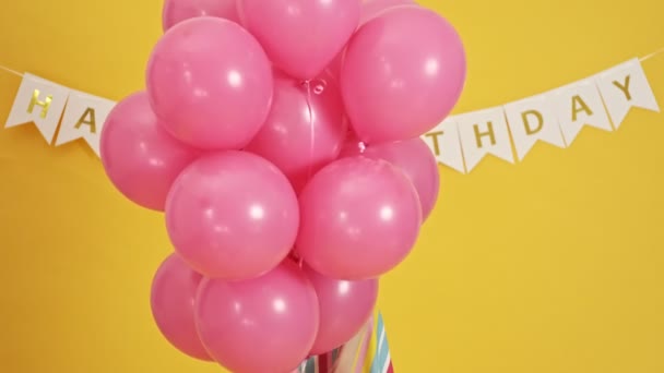 A smiling woman is peeping from pink balloons and waving her hand isolated over a yellow background - Imágenes, Vídeo