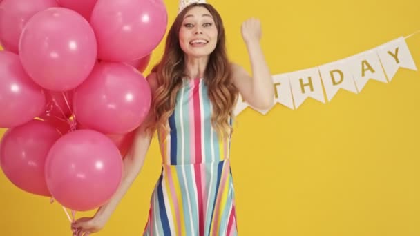 A happy woman is holding pink balloons and doing a winner gesture isolated over a yellow background - Filmati, video