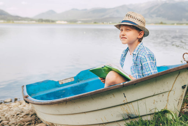 Dreaming boy with book sits in old boat on the mountain lake bank - Photo, image
