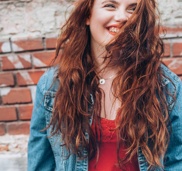Sincerely smiling Red curled hair caucasian teen girl with applied red lipstick lips with blue eyes fashion vertical portrait on the red brick wall background. Natural people beauty concept image. - Foto, Bild
