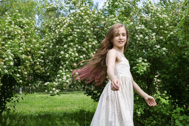 beautiful white girl 11 years old with long hair in a white dress whirls and laughs in a blooming garden - Foto, Bild