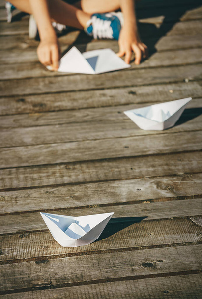 Child makes a paper boats sitting on wooden pier - Foto, Imagen