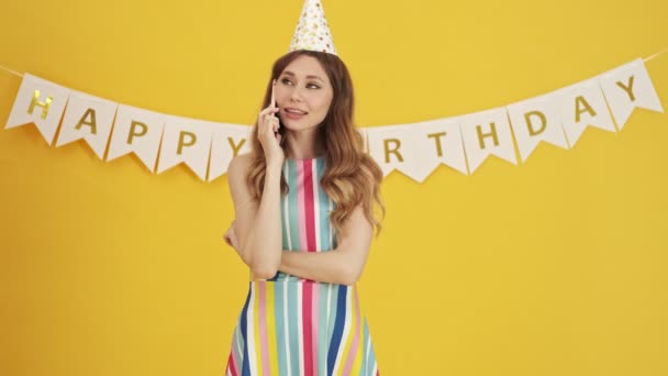 An emotional excited birthday-woman is talking on her smartphone standing isolated over a yellow background - Imágenes, Vídeo