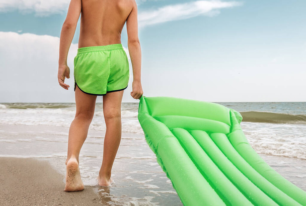 Мальчик с зеленым плавательным матрасом стоит на линии моря
 - Фото, изображение