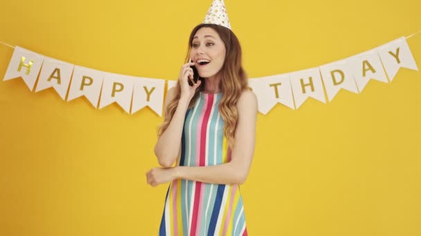 A pleased birthday-woman is talking on her smartphone standing isolated over a yellow background - Πλάνα, βίντεο