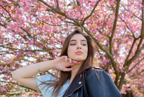 Das junge hübsche Mädchen mit den langen Haaren genießt die Schönheit der Frühlingsnatur in der Nähe des blühenden Sakura-Baumes. Mädchen in Lederjacke in Sakura. - Foto, Bild