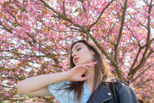 Joven chica hermosa con el pelo largo disfruta de la belleza de la naturaleza de primavera cerca del árbol de sakura en flor. Chica en una chaqueta de cuero en sakura
. - Foto, imagen