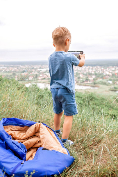 Paikallinen loma. Söpö poika, jolla on lepo makuupussissa vuoren huipulla lähellä kaupunkia ja jutteleminen ystävien kanssa internetin kautta, ulkona kesäviikonloppuna, sosiaalisen etäisyyden elämäntapa - Valokuva, kuva