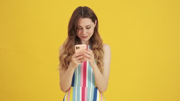 A focused pleased woman is using her smartphone standing isolated over a yellow background - Metraje, vídeo