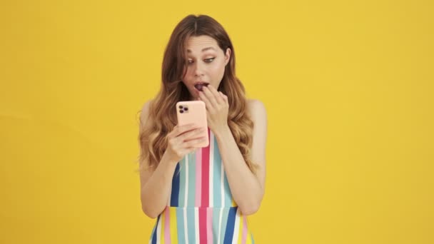 A happy surprised woman is watching something excited on her smartphone standing isolated over a yellow background - Video