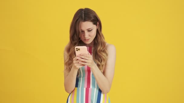 A displeased woman is writing message on her smartphone standing isolated over a yellow background - Imágenes, Vídeo