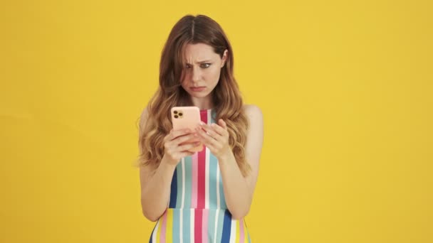 An angry annoyed woman is writing message on her smartphone standing isolated over a yellow background - Imágenes, Vídeo