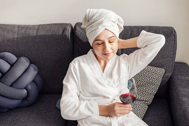 Mujer joven después de la ducha en albornoz con copa de vino tinto sentado en la sala de estar
. - Foto, imagen
