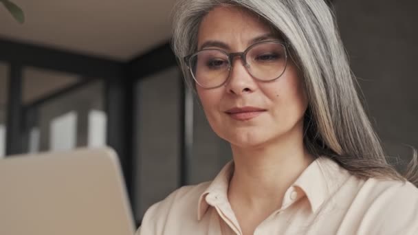 A close-up view of a beautiful mature woman in eyeglasses is looking to her silver laptop computer at home - Video, Çekim