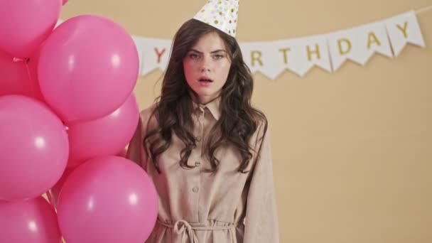 A displeased tired woman in a party cone is holding pink air balloons standing isolated over a beige background - Imágenes, Vídeo