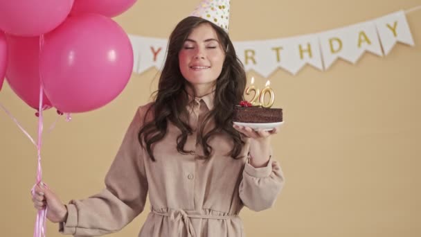 A smiling pleased woman in a party cone is holding pink air balloons and a birthday cake while making a wish and blowing candles isolated over a beige background - Imágenes, Vídeo