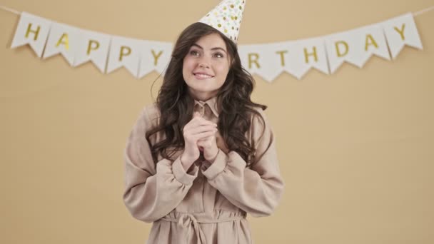 A lovely birthday-woman in a party cone is thinking about something while making a wish standing over a beige background - Video