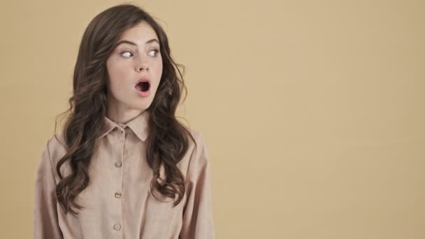 A shocked surprised woman is pointing to the free space isolated over a beige background - Video