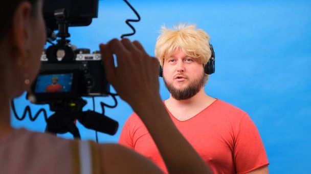 hombre gordo raro en una peluca y una camiseta rosa hace un video blog sobre un fondo azul
. - Foto, Imagen