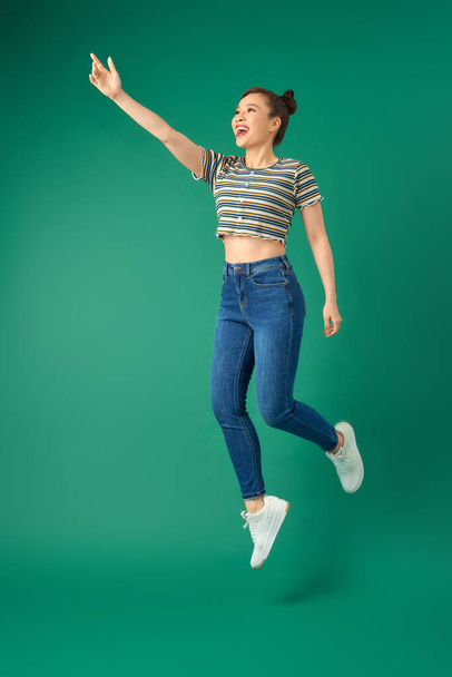 Full length of cheerful young Asian woman or teenage girl jumping in air over green background. - Foto, imagen