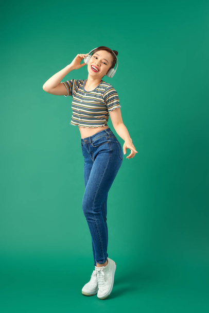 Full length portrait of joyful Asian woman on casual clothing dancing and listening to music with headphone over green background. - Foto, Imagen