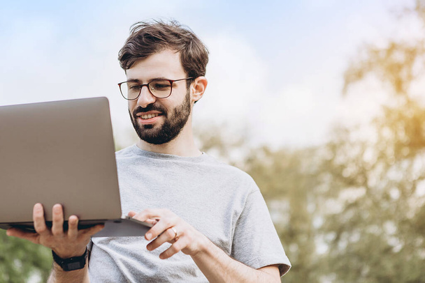 Attractive young man with dark hair in glasses works outdoors with a laptop on a sunny day. Bearded hipster guy wears casual clothes and works remotely at a quarantined company - Φωτογραφία, εικόνα