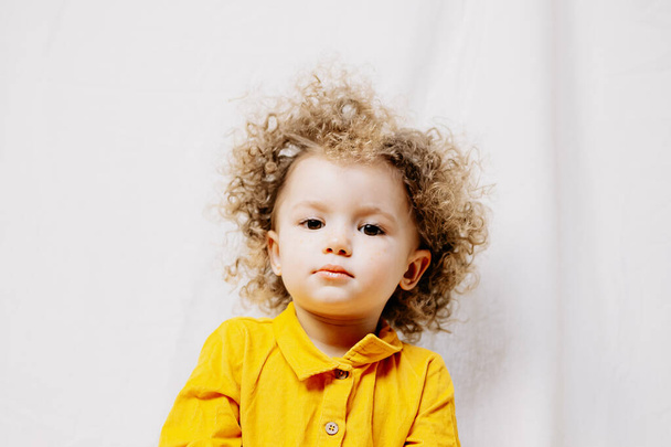 Portrét atraktivní roztomilé dítě oblečené ve žlutých šatech pózovat na světlém pozadí v domácnosti improvizované foto studio - Fotografie, Obrázek