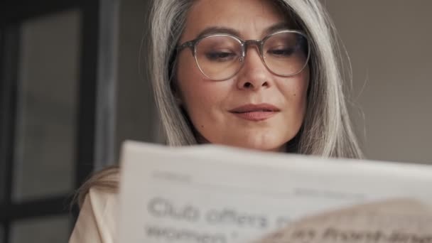 A close-up view of a beautiful mature woman wearing eyeglasses is reading a newspaper sitting at the table at home in the morning - Materiał filmowy, wideo