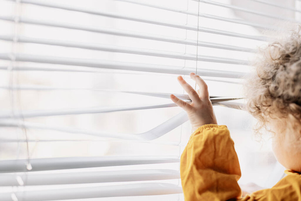 The child opens the blinds with his hands and looks out the window outside. Copy space - Foto, Imagem