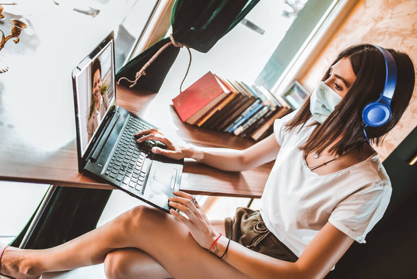 Casual kobieta freelancer w masce twarzy, słuchawki za pomocą laptopa do zdalnej pracy online w kawiarni, rozmowy wideo. - Zdjęcie, obraz