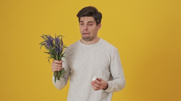 Unhappy sick man sniffing flowers then having allergy and using spray after that over yellow background - Imágenes, Vídeo