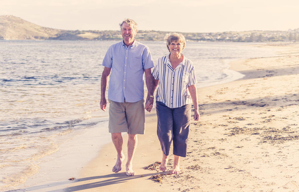 Happy senior couple embracing and holding hands walking on empty beach at sunset. Active healthy elderly woman and man on a romantic walk by the sea. Aging together and retirement lifestyle concept. - Foto, Imagem