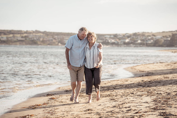 Happy senior couple embracing and holding hands walking on empty beach at sunset. Active healthy elderly woman and man on a romantic walk by the sea. Aging together and retirement lifestyle concept. - Photo, image