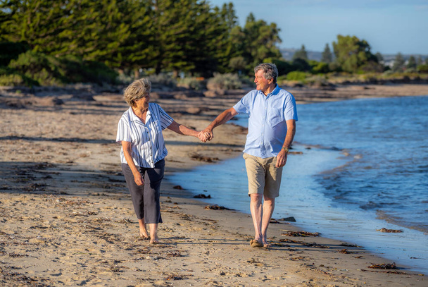 Happy senior couple embracing and holding hands walking on empty beach at sunset. Active healthy elderly woman and man on a romantic walk by the sea. Aging together and retirement lifestyle concept. - Photo, Image