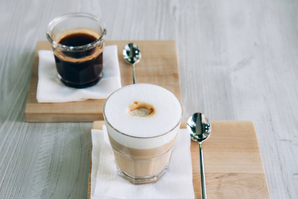 Fekete kávé és Cappuccino üvegpohárban, fából készült tálcán. Reggeli americano habbal a fa asztalon. - Fotó, kép