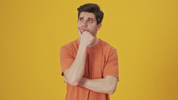 Pensive handsome man wearing in t-shirt thinking about something while holding his chin over yellow background - Filmagem, Vídeo