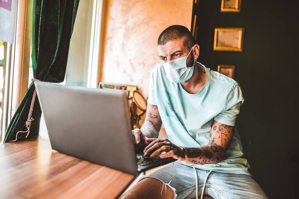 Casual tatuaje hombre freelancer en mascarilla facial, utilizando el ordenador portátil para trabajar de forma remota en línea en la cafetería - Foto, imagen