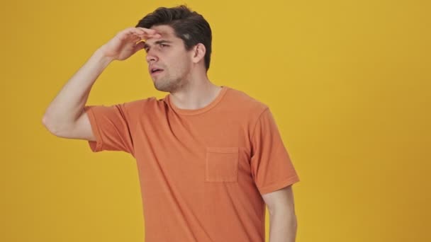 Concentrated handsome man wearing in t-shirt looking around with hand on forehead then becoming happy over yellow background - Felvétel, videó
