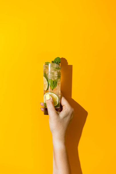 Nainen käsi pidä lasia vesipisaroita Mojito cocktail juoda lime Ice ja minttu kirkkaan keltaisella pohjalla. Summer Art ruoka käsite keltaisella pohjalla. - Valokuva, kuva
